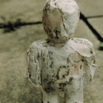Kleine weiße Tonfigur sitzt mit angezogenen Knien auf Betonboden | Small white clay figure sitting with knees drawn up on concrete floor