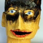 Gelbe Skulptur eines Kopfes mit blauen Augen, schwarzen Farbspuren und rotem Mund | Yellow head sculpture with blue eyes, black paint drips, and red open mouth