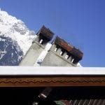 Zwei Schornsteine auf schneebedecktem Dach vor verschneitem Alpengipfel | Two chimneys on snow-covered roof against snowy Alpine peak