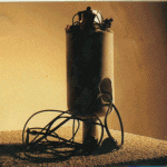 Zylindrisches Metallgerät mit Kabeln auf Betonboden vor gelber Wand | Cylindrical metal device with cables on concrete floor against yellow wall