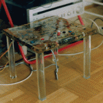 Kleiner Metalltisch mit farbig bemalter Platte und Elektronikkomponenten, Kabel auf Parkettboden | Small metal table with colorfully painted top and electronic components, cables on parquet floor
