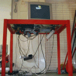 Roter Metallrahmen mit freiliegenden PC-Komponenten und Kabeln, darüber CRT-Monitor | Red metal frame with exposed PC components and cables, CRT monitor on top