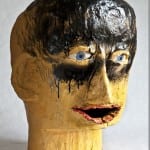 Skulptur eines Kopfes, goldgelb, schwarze Farbe läuft über Gesicht, blaue Augen | Sculpted head, gold-yellow, black paint dripping over face, blue eyes, open mouth