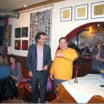 Zwei Männer stehen lachend in einem Kunstcafé mit bunten Gemälden an den Wänden | Two men laughing in an art café with colorful paintings on the walls