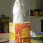 Weihnachtsmann-Figur in transparenter Cola-Mix-PET-Flasche auf blauem Tisch | Santa Claus figurine inside a transparent Cola-Mix PET bottle on blue table