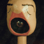 Bemalter Holzkopf auf Sockel mit geschlossenen Augen und großem offenem Mund als Linse | Painted wooden head on base with closed eyes and large open mouth housing a lens
