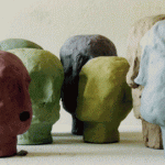 Sieben grob geformte Tonköpfe in verschiedenen Farben auf hellem Untergrund | Seven roughly shaped clay heads in various colors on a light surface