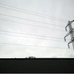 Strommast mit Hochspannungsleitungen vor grauem Himmel, schneebedeckte Berge | Power pylon with high-voltage lines against grey sky, snow-capped mountains