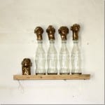 Vier Coca-Cola-Flaschen mit bronzenen Menschenköpfen auf Holzregal | Four Coca-Cola bottles topped with bronze human heads on a wooden shelf