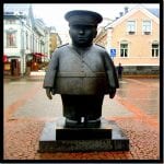 Rundliche Bronzestatue eines Polizisten mit Mütze auf gepflastertem Stadtplatz | Chubby bronze statue of a police officer with cap on a cobblestone town square