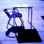 Leerer Café-Stuhl und Metalltisch mit Aschenbecher, blau verfärbt, starker Schatten | Empty café chair and metal table with ashtray, blue tinted, strong shadow cast