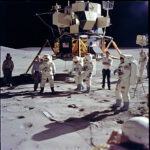 Astronauten in weißen Raumanzügen vor Mondlandefähre mit US-Flagge auf grauem Mondboden | Astronauts in white spacesuits before lunar lander with US flag on grey moon surface