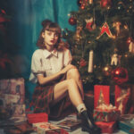 Junge Frau in weißer Bluse sitzt vor geschmücktem Weihnachtsbaum mit Geschenken | Young woman in white blouse sitting before decorated Christmas tree surrounded by gifts