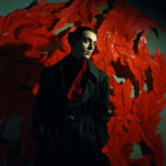 Person in schwarzem Mantel mit rotem Schal vor leuchtend roten Blättern | Person in black coat with red scarf against vivid red sculptural leaves